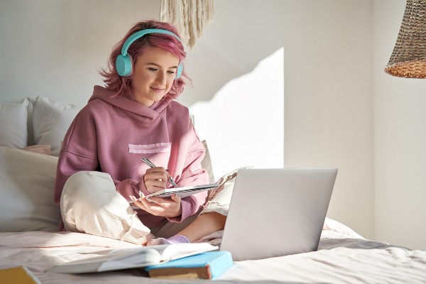 A girl with pink hair and blue headphones sitting on her bed with a Chromebook and writing notes.